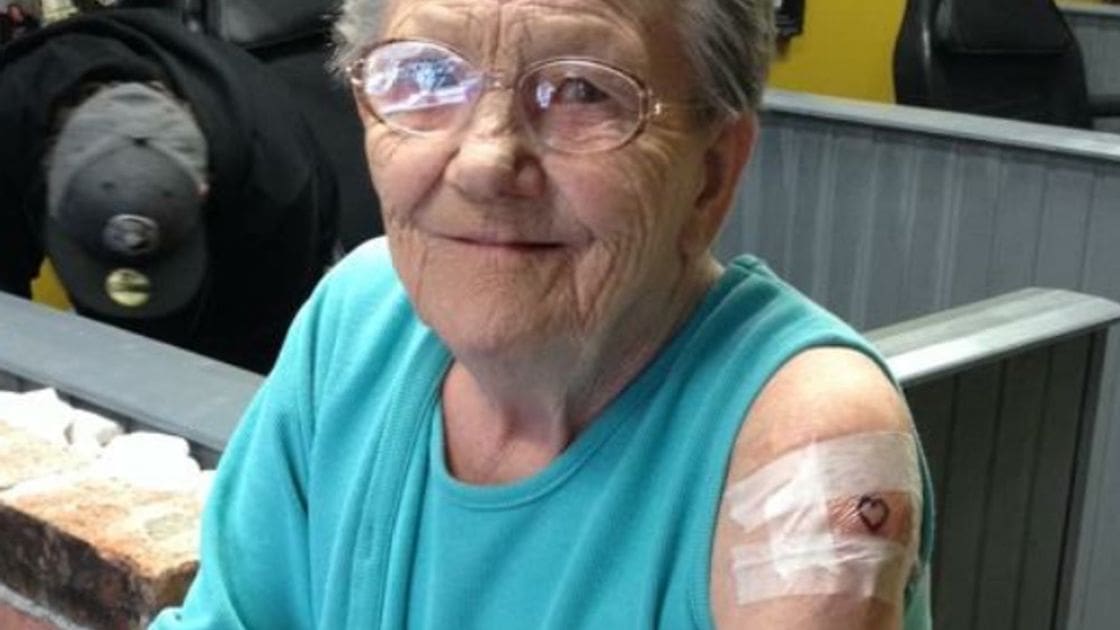 An older adult female wearing glasses with thin frames, a sleeveless teal shirt, and a bandage with a heart symbol on her upper arm, standing outdoors in what appears to be a public space during daytime.
