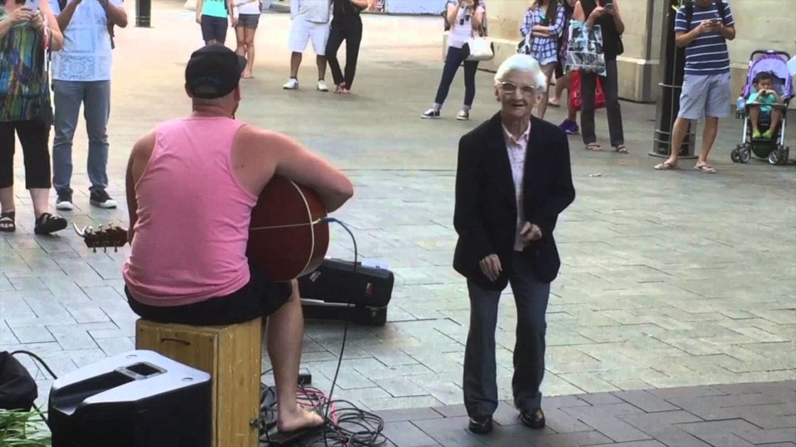 An urban plaza scene with a street musician playing guitar, an older woman standing nearby, and various people in the background; an amplifier and suitcase are near the musician, stroller visible in the distance; architectural style suggests European setting