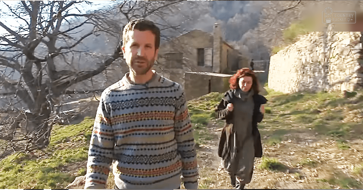 Middle-aged man wearing a patterned sweater stands in an old village setting with smoke rising from one of the stone buildings; season is likely late autumn or early spring, light suggests midday.