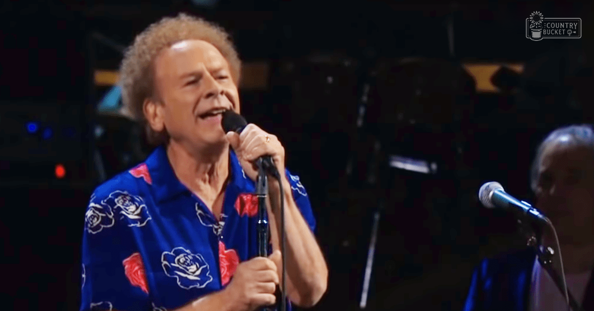 A middle-aged man wearing a loose-fitting blue Hawaiian-style shirt with red and white floral patterns performs on stage at an indoor concert venue under bright stage lighting.
