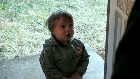 A toddler wearing an olive green hooded sweatshirt outdoors near a doorway with concrete ground and grass visible in the background.