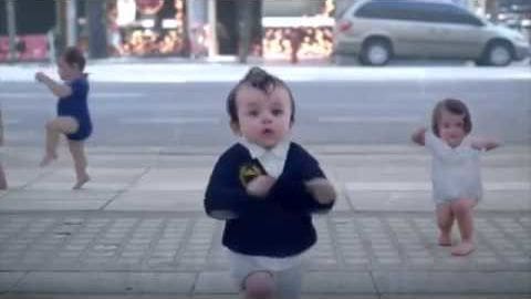A toddler in an urban plaza with a white minivan visible in the background, wearing a dark blue vest over a white shirt and light-colored shorts or pants. A female infant/toddler in a blue one-piece outfit is seen on the left, while another female infant/toddler in a white onesie/dress appears to the right.
