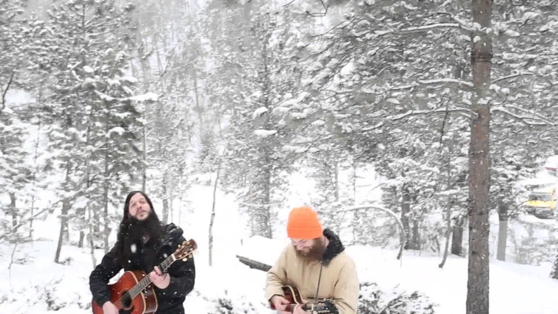 A snowy forest scene with two adult males performing a musical act, one in black and the other in orange, under diffused winter light.
