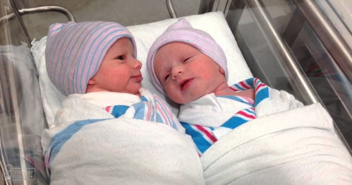 Two newborn babies wearing striped knit hats in a hospital bassinet, clinical lighting illuminating their white blankets and pink-blue stripes.