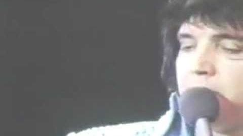 A young adult male singer performing at a concert, wearing a white patterned dress shirt with stage lighting and dark background.
