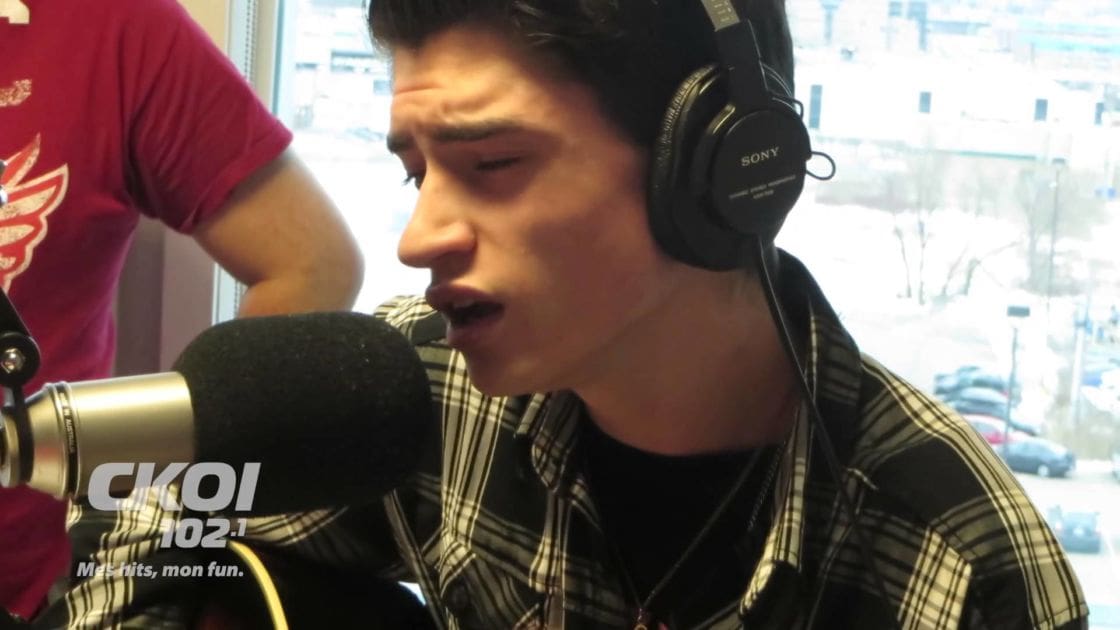 A young adult male wearing a plaid shirt with large Sony headphones, standing in a radio studio during winter with natural daylight and snow visible outside.