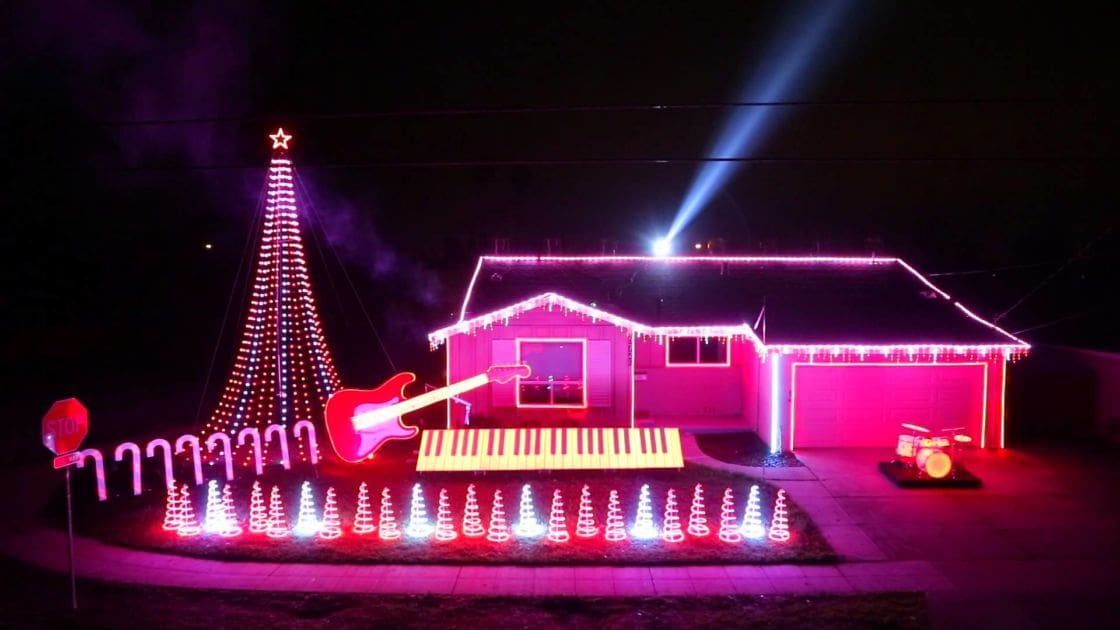 A decorated urban residential area with Christmas lights, a pink electric guitar, yellow keyboard, drum set, stop sign, and string lights forming musical notes on the ground at night during the holiday season.