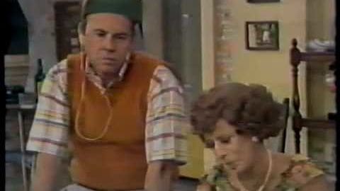 A middle-aged adult male wearing an orange vest and striped shirt, accompanied by a child in a light-colored floral top. The setting is an indoor residential space with a staircase and framed artwork.