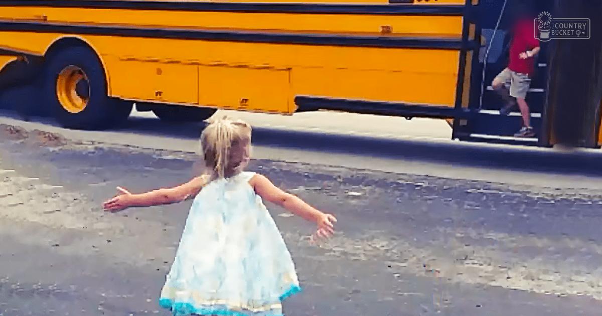 A toddler in a light blue floral dress stands near an adult male wearing red and khaki, with a yellow school bus in the background at what appears to be a school or transportation facility on a warm day.