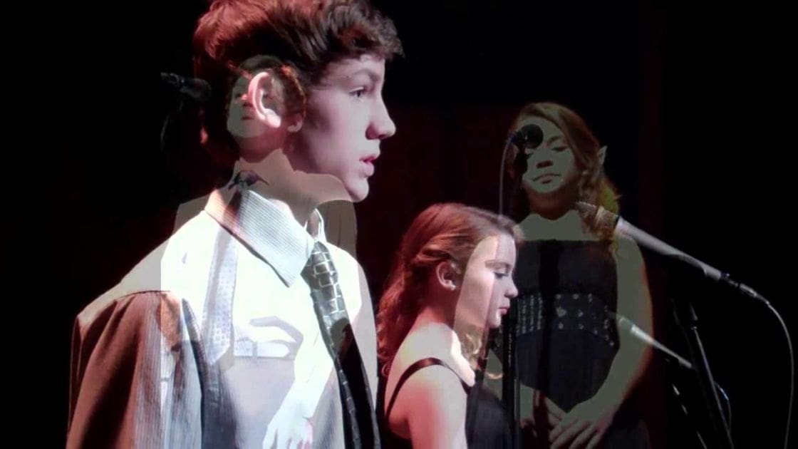 A young adult male singer in a white shirt with a patterned tie, brown jacket, and light brown hair performing on stage.