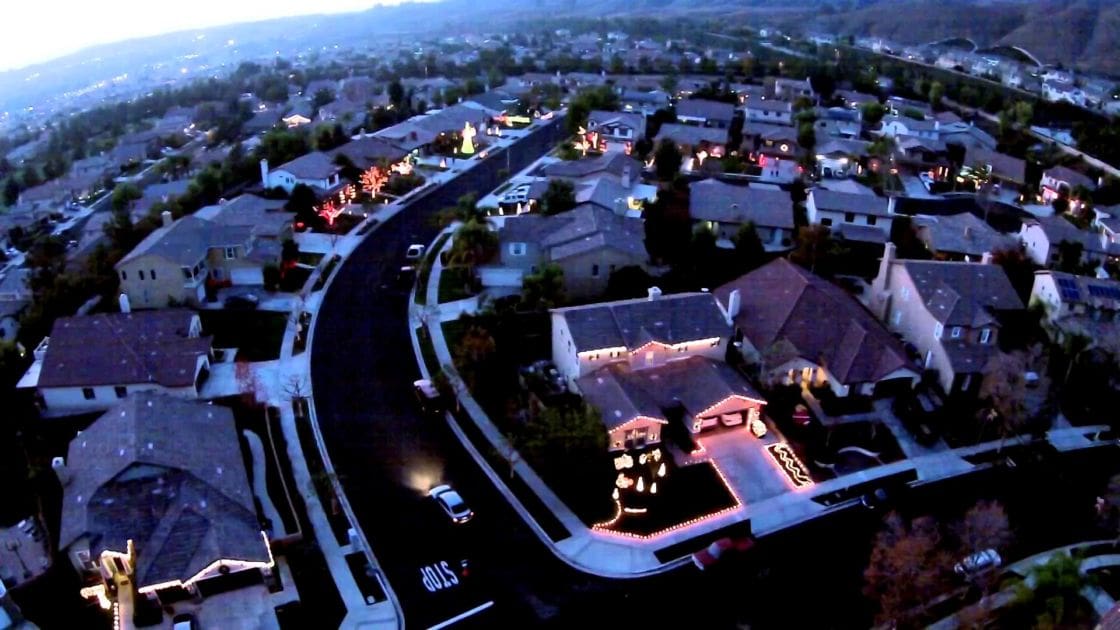 A residential neighborhood at dusk, with houses adorned for Halloween with muted blue and gray tones in the sky and buildings, accented by bright white Halloween lights on one house.