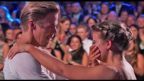 A couple dancing on a stage in front of an audience during what appears to be a dance competition or performance, with artificial stage lighting illuminating the dancers and a mix of white and gray clothing visible.