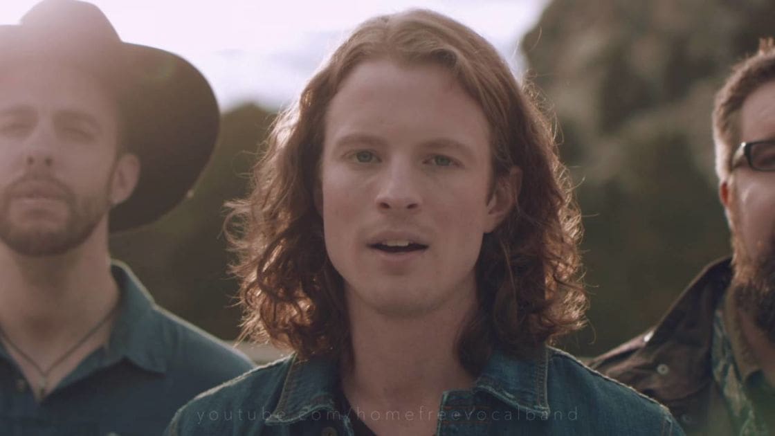 A young adult male wearing a denim jacket outdoors in a natural setting with other adults in camouflage shirts nearby.