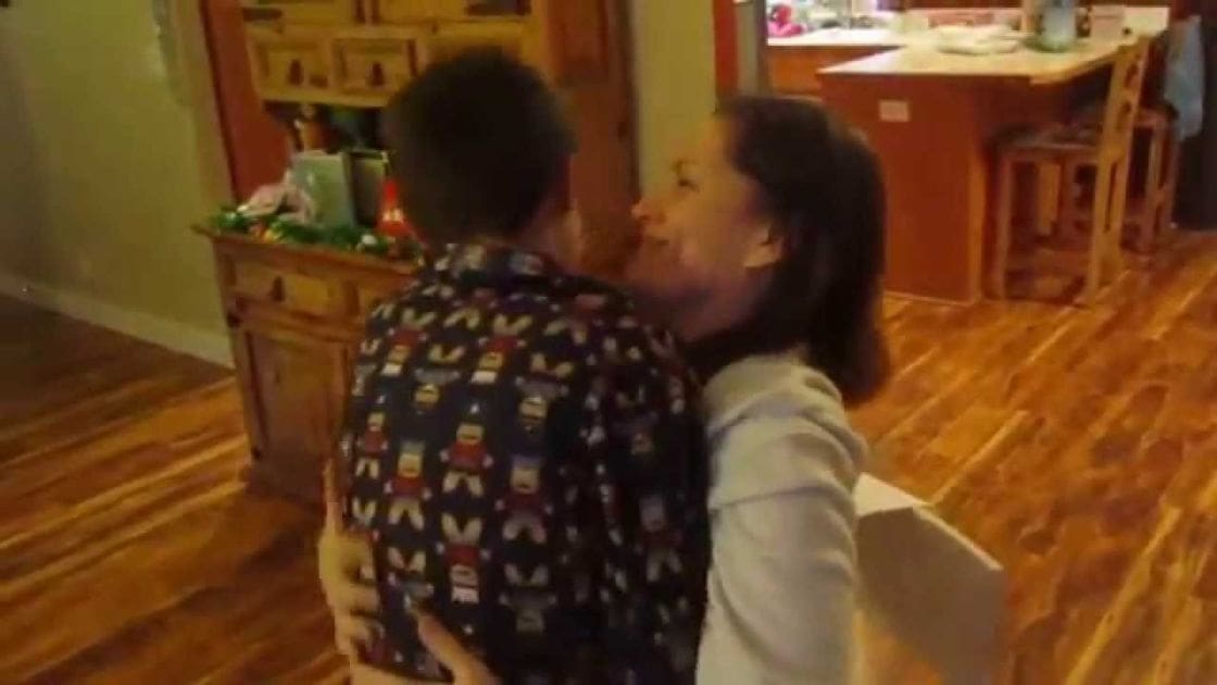 A toddler wearing a dark shirt with 'DINOSAUR' text and cartoon characters, held by an adult female in a home setting with wooden cabinets and kitchen elements in the background.