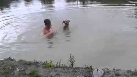 An adult male swimmer with a medium-sized brown dog by a body of water, vegetation visible at the shoreline in spring or summer.