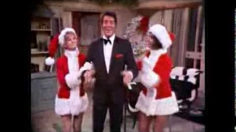 Adult male in a black tuxedo at an outdoor Christmas-themed event with young children in red and white Santa outfits holding something.
