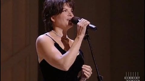 Middle-aged woman singing into a microphone at an indoor event, wearing a black sleeveless top with thin straps and a low neckline.