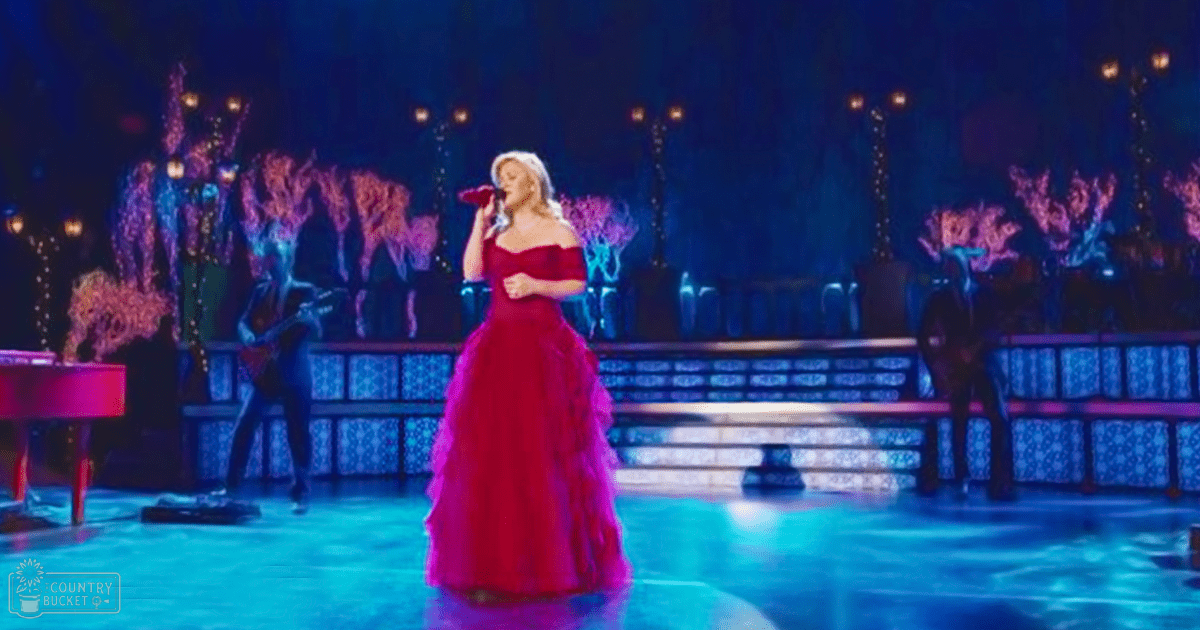 A female adult singer in a red off-the-shoulder gown with ruffled details performs on an indoor stage during what appears to be an evening concert.