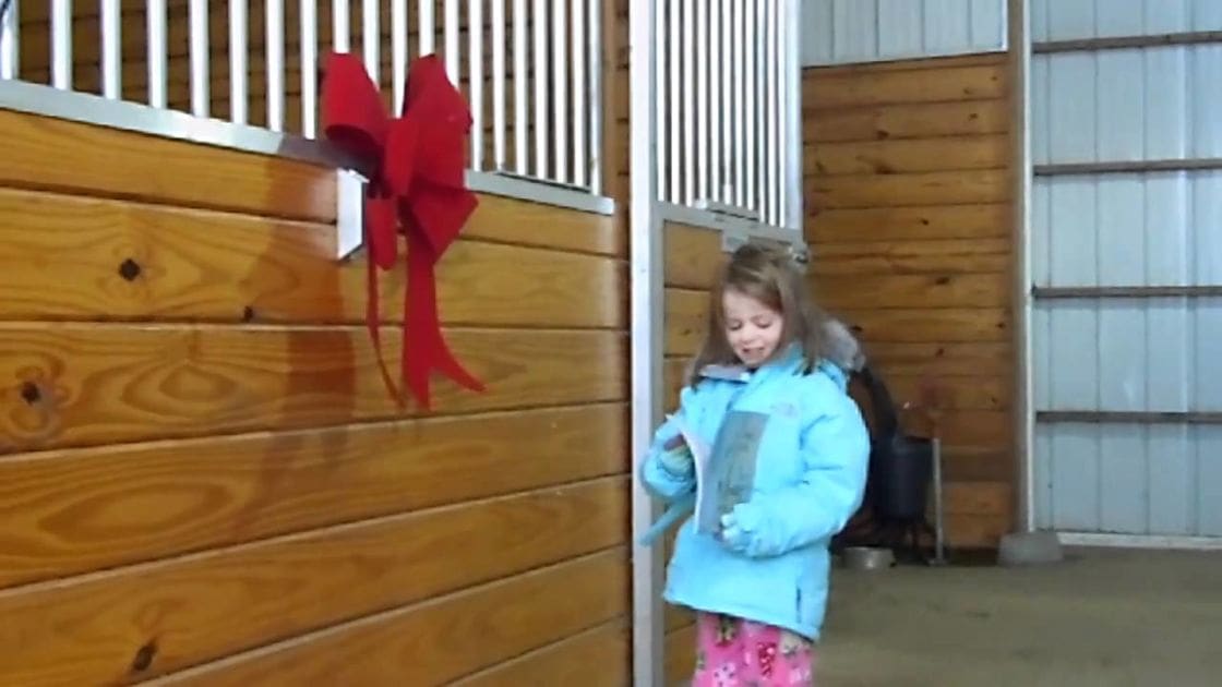A young child in a light blue puffer jacket with a hood, wearing pink patterned pajama-style pants inside an indoor stable setting adorned with a red decorative bow on the wall.