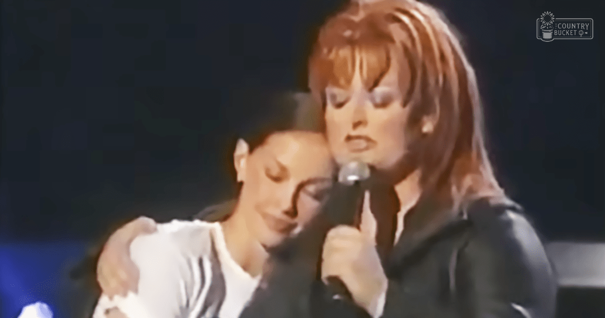 A musical performance scene with two women, one wearing an athletic top and another in formal attire, under dim stage lighting.