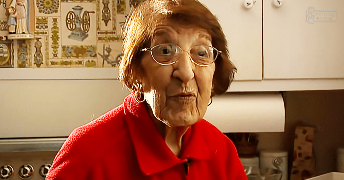 Older adult female in a kitchen setting with red jacket, round glasses, hoop earrings; white cabinets, decorative tiles, stove knobs visible.