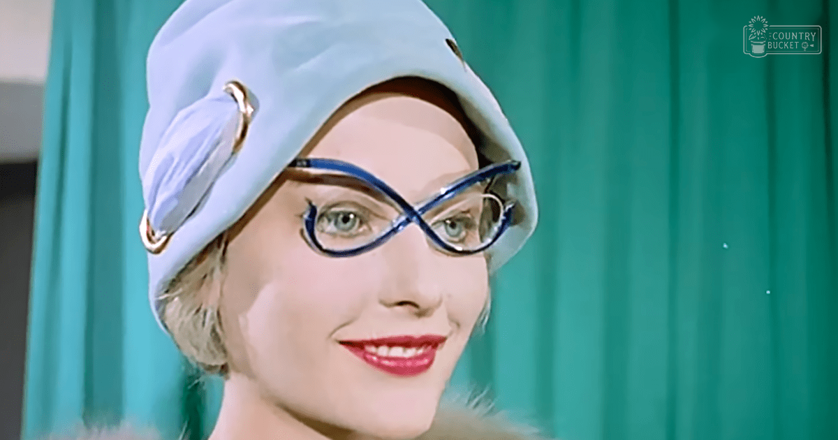 An adult female wearing a light blue beret with decorative gold trim, large dark blue glasses, in an indoor setting with teal curtains.