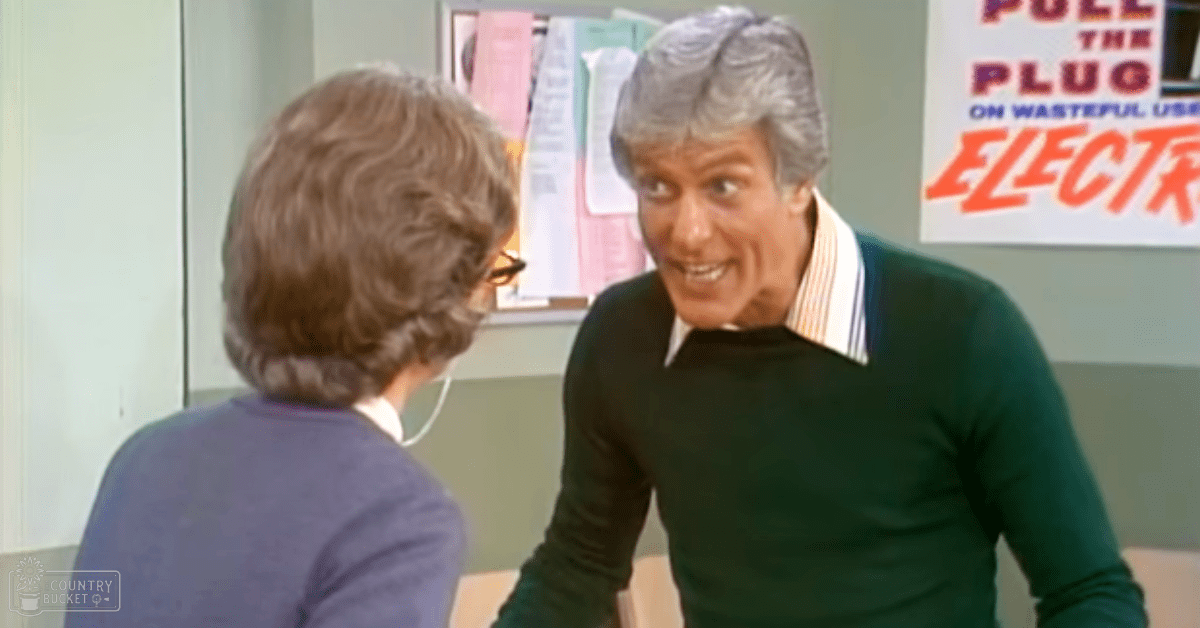 A middle-aged man wearing a dark green long-sleeved shirt with white and orange stripes at the collar, paired with an older adult female in a purple top with large hoop earrings. They are indoors, possibly in an office or classroom setting.