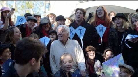 A diverse group celebrates outdoors under natural light in an urban park setting, holding signs and flags, suggesting a birth-related gathering.