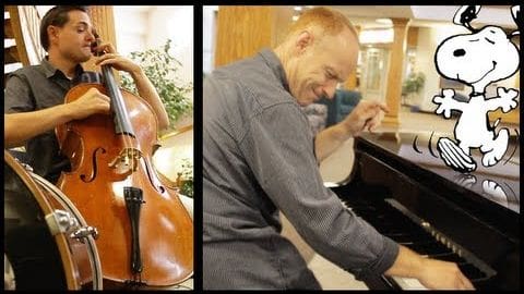 An adult male singer wearing a dark gray button-up shirt and black pants, accompanied by another adult male in a similar outfit. The setting is an indoor performance space with a polished cello on the left and a piano on the right, surrounded by natural light from windows.