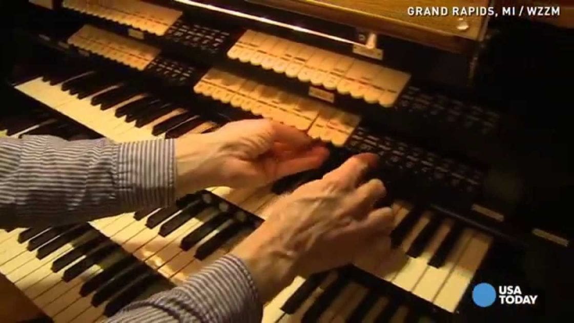 An older adult male singer playing an organ indoors with artificial lighting, wearing a striped shirt from the mid-20th century.
