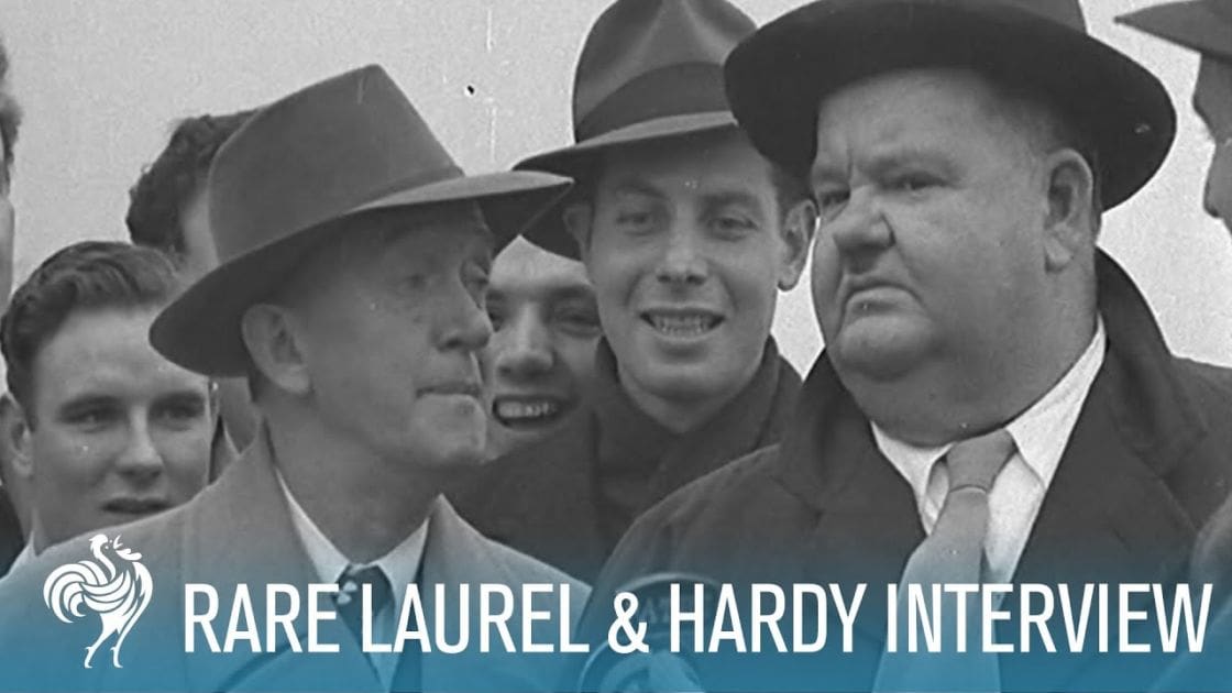 An adult male singer in a fedora-style hat and dark-colored suit jacket over a white collared shirt and tie during what appears to be an interview with Laurel & Hardy's era theme, held outdoors under overcast lighting.
