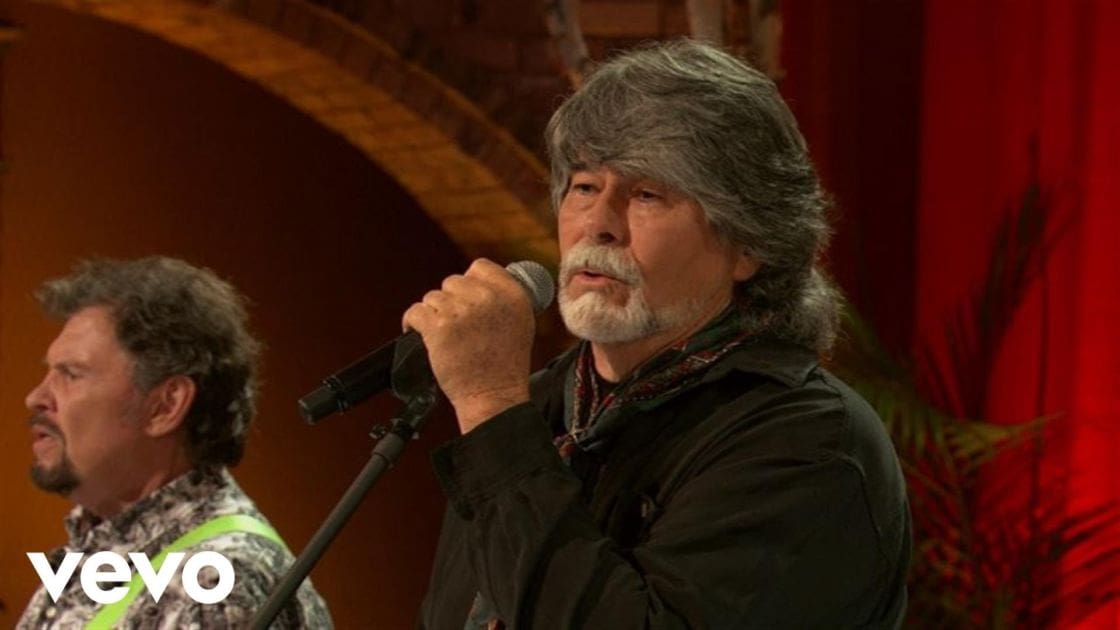 An older adult male wearing a dark green jacket and patterned scarf sings into a microphone at an indoor performance venue with red curtains.