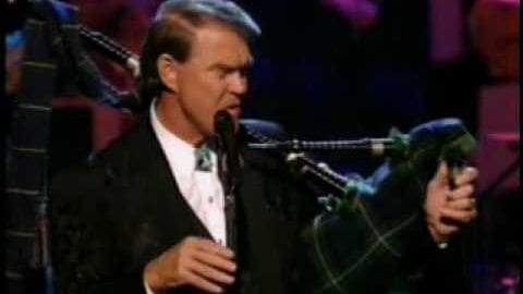 A middle-aged male singer performing at an indoor event, wearing a dark blazer over a white patterned tie shirt, holding a microphone and bagpipe.