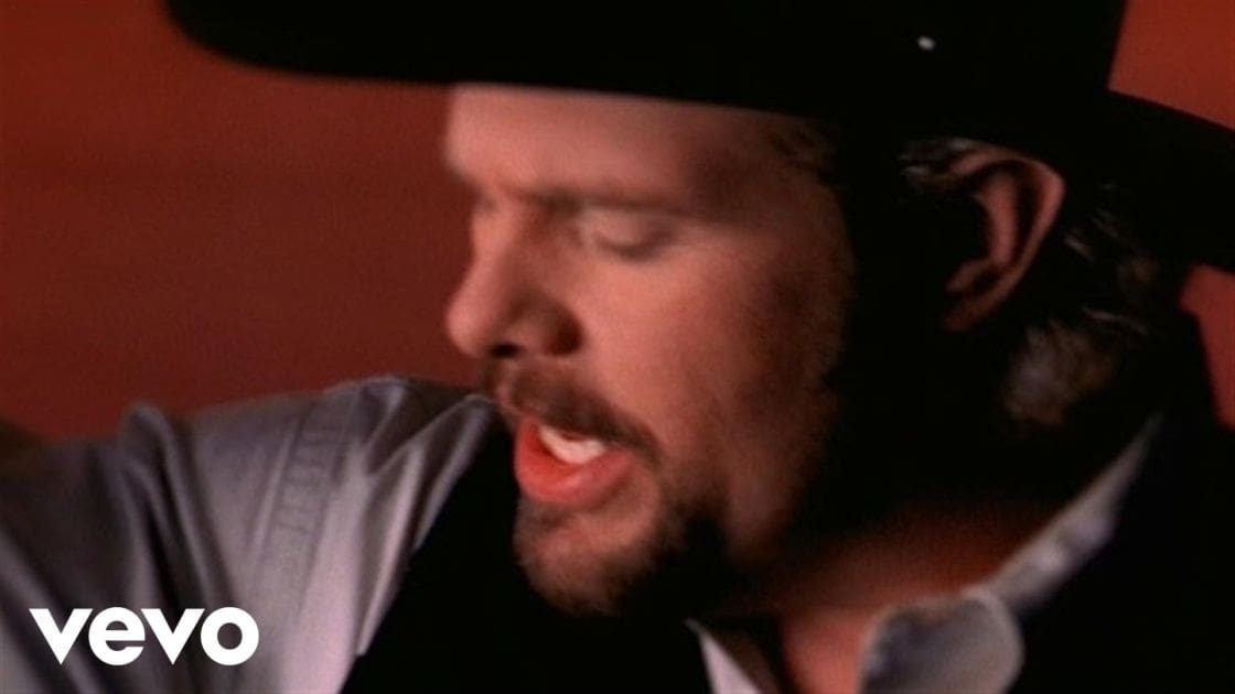 An adult male singer wearing a collared shirt and vest with a subtle striped pattern, accompanied by a black cowboy hat, in an indoor setting with warm lighting.