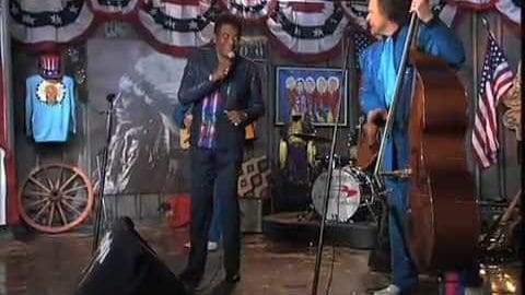 Adult male singer performing with a double bass player in an indoor venue, possibly a bar or small concert hall. The setting is decorated with American flags and patriotic themes.