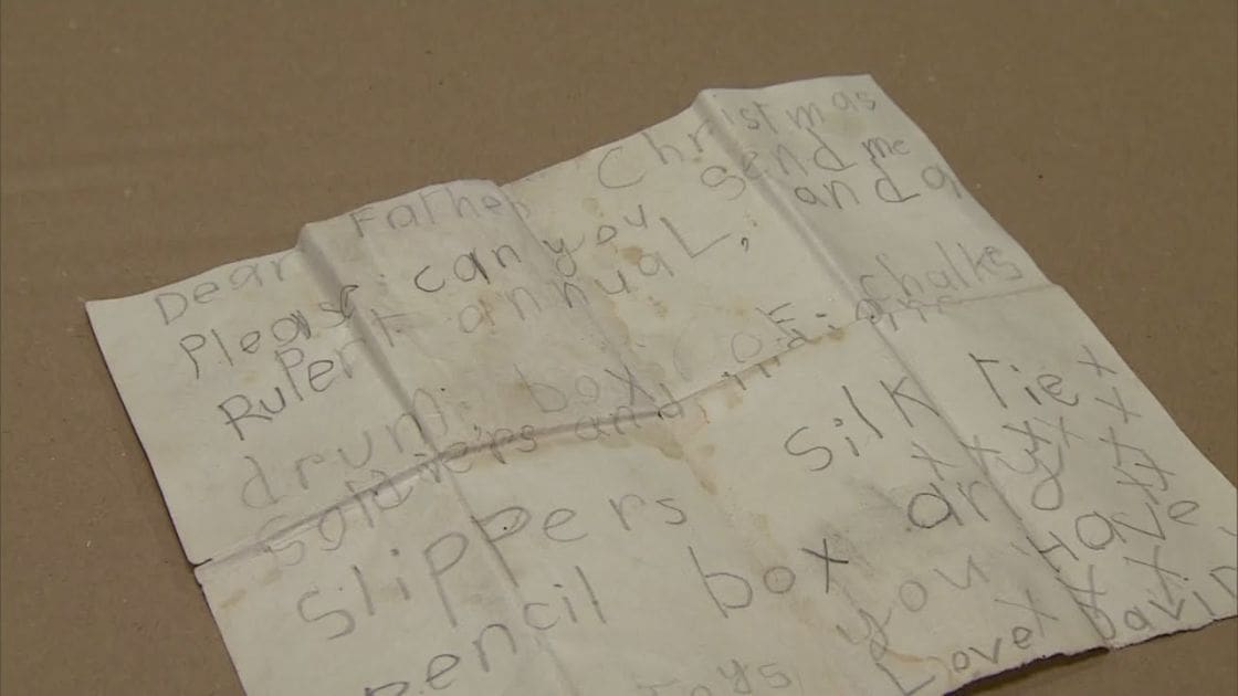 An old, crumpled letter addressed to Father Christmas with a wish list for gifts on a cardboard surface indoors.