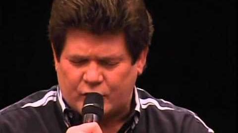 A middle-aged adult male sings into a microphone in an indoor setting with dark background lighting, possibly a stage or performance space.