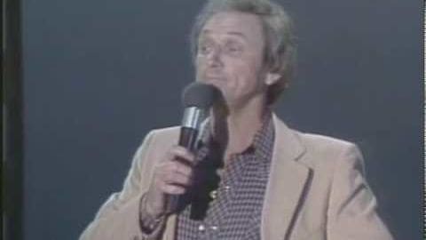 A middle-aged adult male wearing a beige blazer over a checkered shirt indoors, holding a handheld dynamic microphone in front of a plain dark backdrop.