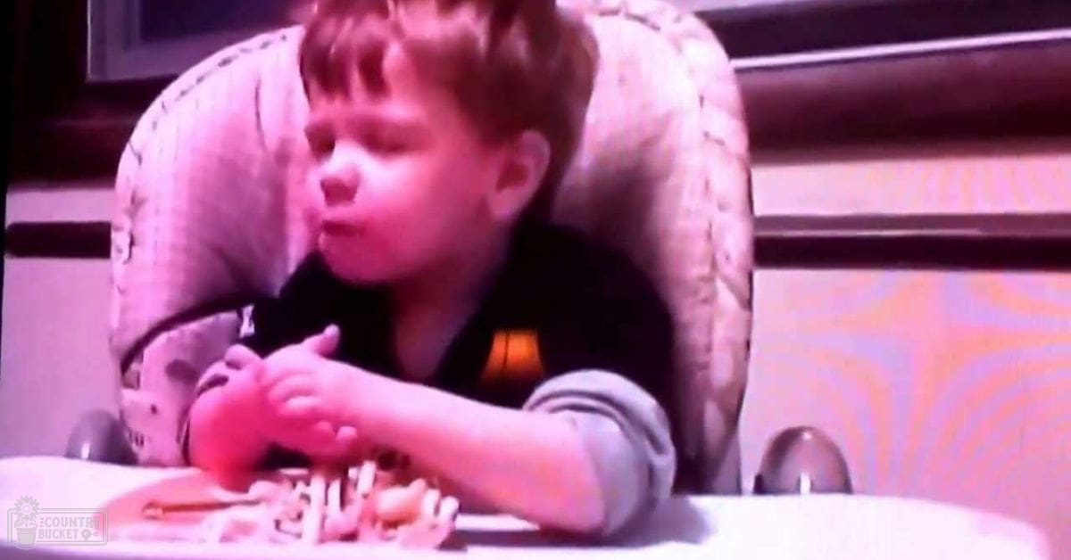 A toddler in a high chair indoors, wearing a dark shirt; food items like pasta are visible on a plate.