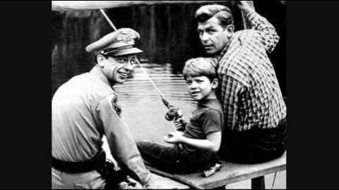Child fishing with adults in a park setting; muted black-and-white photo with soft lighting, outdoor environment, trees or foliage in background.