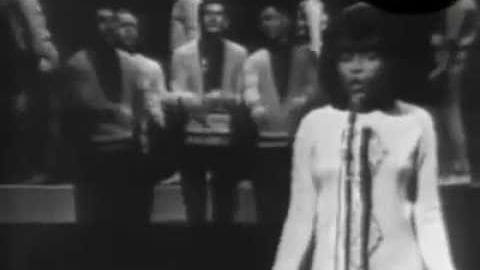 A young adult female stands on a stage or performance setting wearing a loose-fitting white top with long sleeves and a tie-like detail around the neck, surrounded by adults in dark suits. The scene is indoors with focused lighting typical of a stage performance.