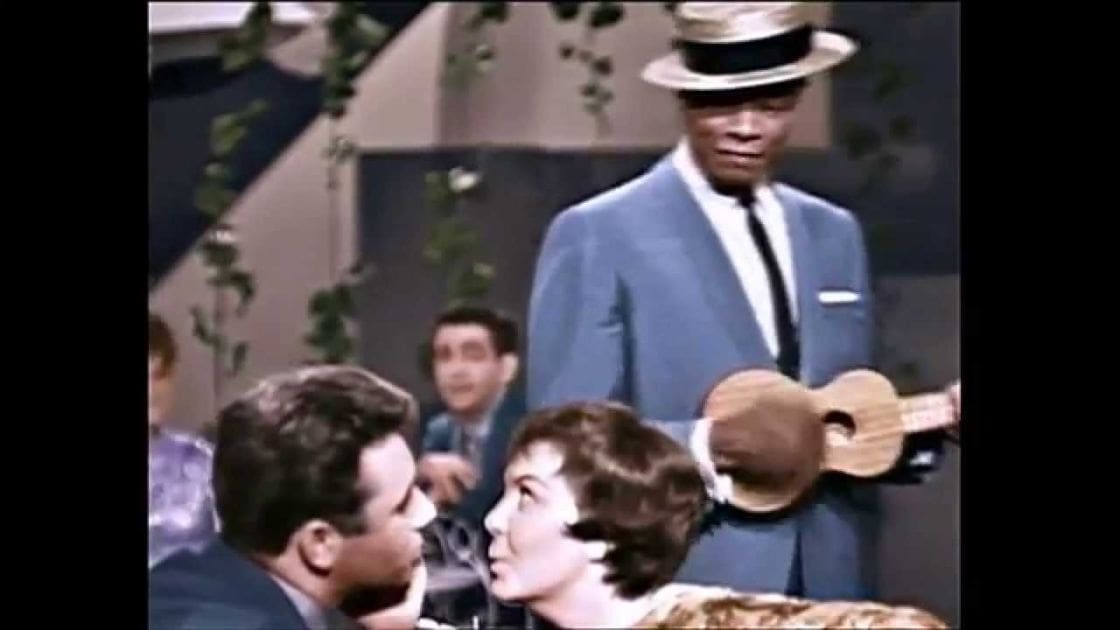 An indoor mid-20th-century event with people in formal attire, decorative greenery hanging on the wall behind them.