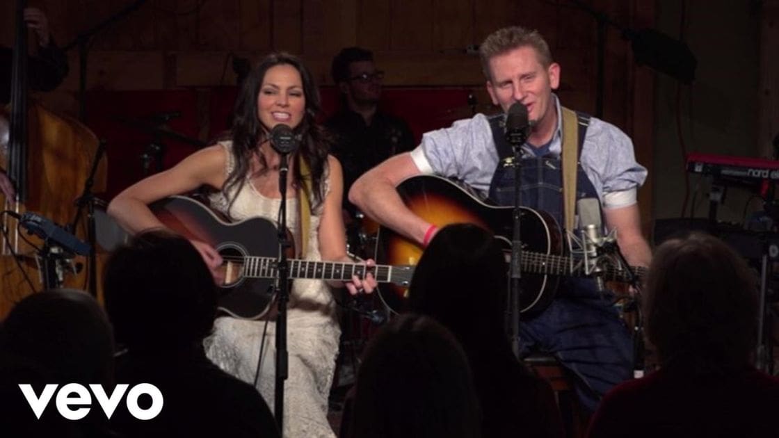 Adult performers in a musical performance setting with microphones, stringed instruments, and wooden walls at night.