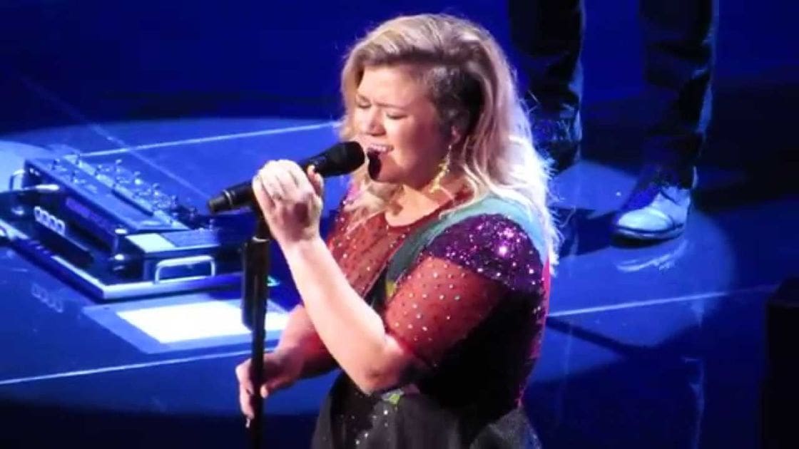 A middle-aged female performer in a red, dotted dress with short sleeves and dark green shoulder straps sings on stage at an indoor concert venue under dramatic blue lighting.