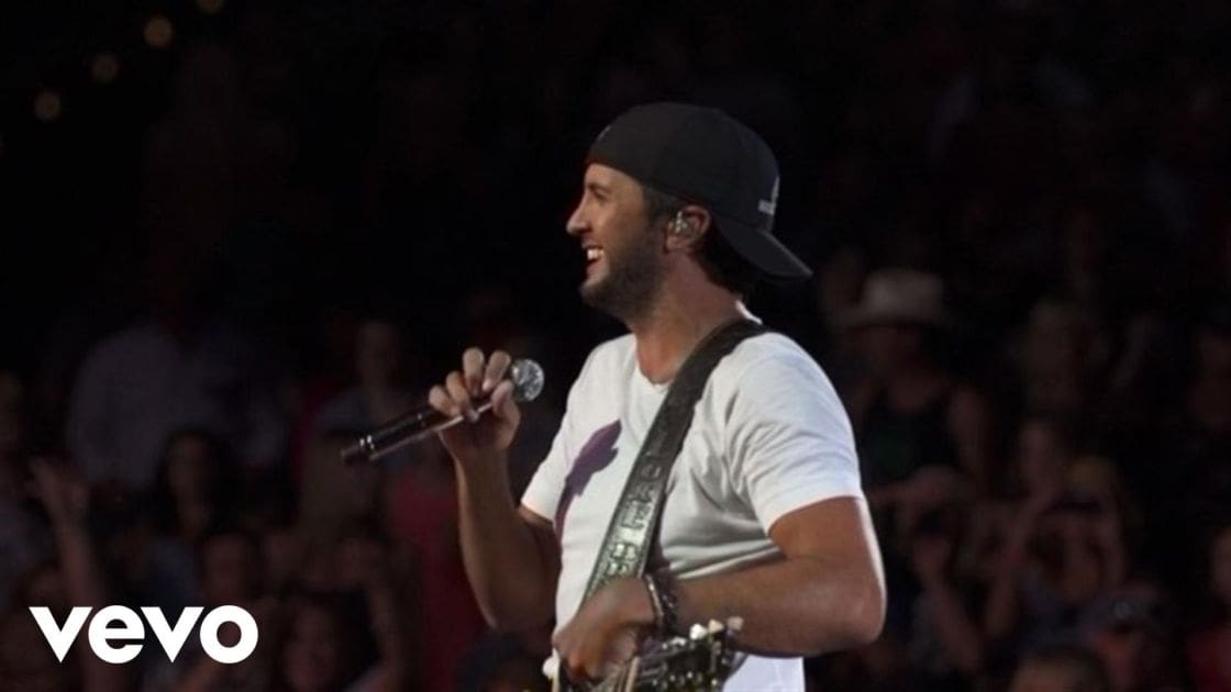 Adult male performer in white short-sleeved T-shirt with purple graphic, black baseball cap backward, holding microphone and guitar strap; stage lighting at indoor concert venue.