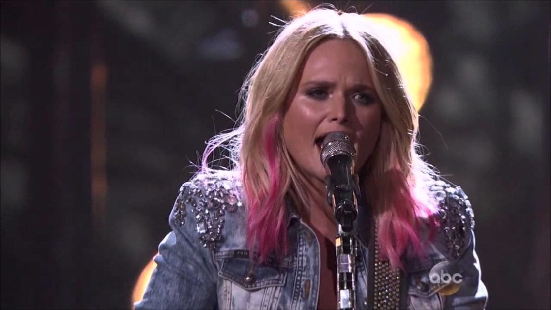 A young adult female performs on stage, wearing a denim jacket with studded embellishments at an awards show.