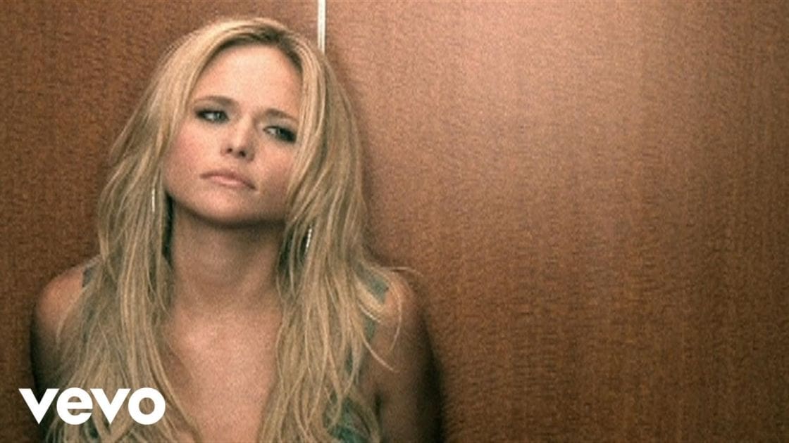 A young adult female wearing a loose-fitting strapless top or dress with a floral green and white pattern in an indoor space with warm lighting.