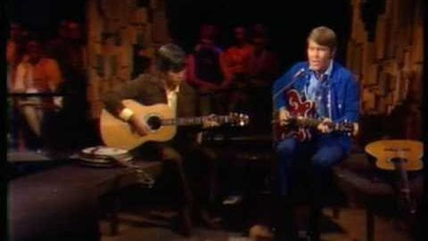 Two young adult male musicians in an indoor performance space, one wearing a dark patterned shirt and blue jeans playing guitar, another in a denim jacket over a white 'FROGS' shirt. The setting is stage-like with decorative elements resembling buildings.