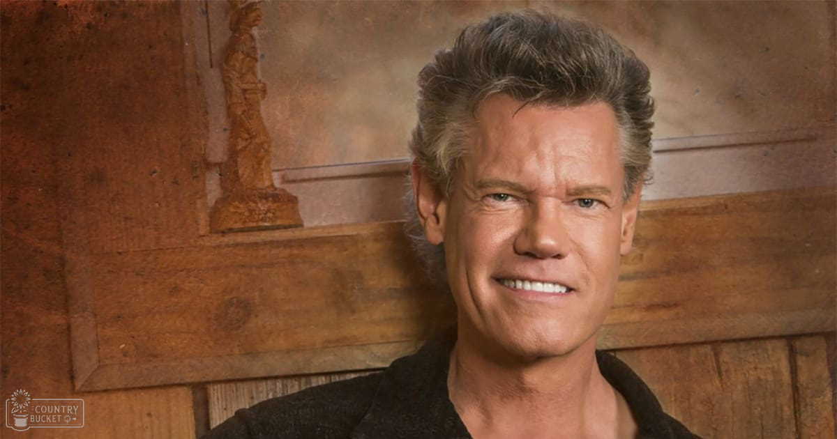 A middle-aged adult male wearing a relaxed fit, dark-colored V-neck sweater in an indoor setting with wooden paneling and a decorative statue-like figure.