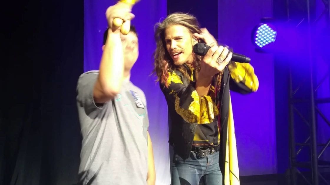 Young adult male singer wearing a gray t-shirt with a logo, performing in an indoor stage setting with another middle-aged man in the background holding a microphone and wearing a black jacket over a yellow patterned shirt. The backdrop is dark with stage lighting.
